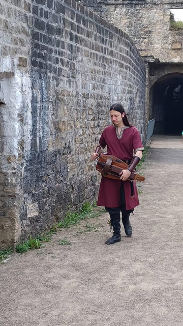 Me in a medieval costume, with a red tunic and black (vegan) leather boots, with (not vegan) leather arm bands, playing my hurdy-gurdy among the stone walls of a castle ruin in northern France.