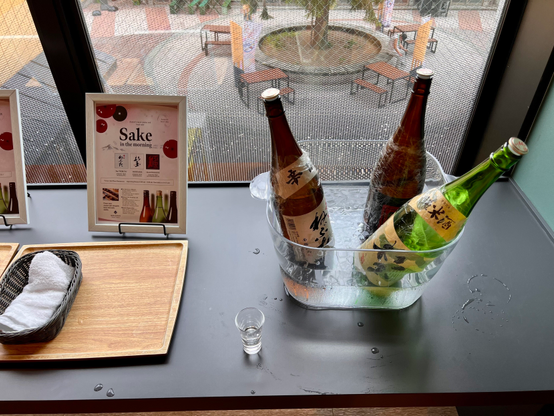 A table displays three bottles of sake in an ice bucket, accompanied by a small glass and a basket with towels. Framed menus about sake are also present, with a view of an outdoor space through a window.