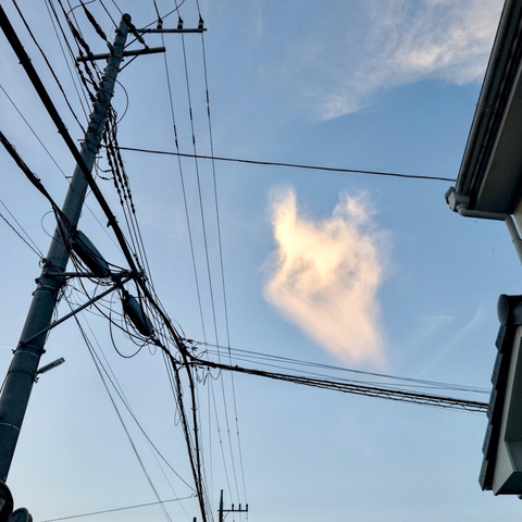 A pole carrying a cobweb of power lines and the outline of a house gable frame a solitary blob of glowing cloud. Blue sky, sunset tones catch the cloud. Whispy edges suggest wind dragging and distorting the cloud’s shape.