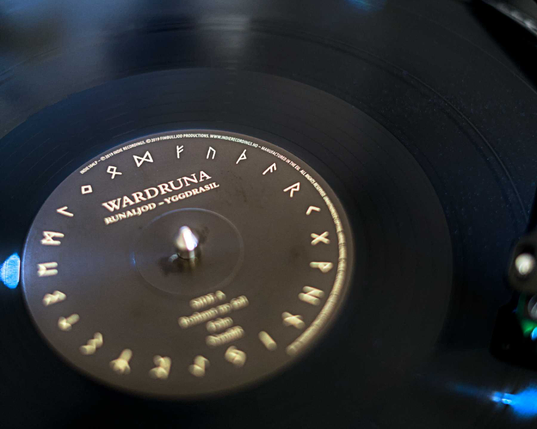 Die schwarze Scheibe einer Schallplatte liegt auf einem Plattenteller. Der Blickwinkel ist auf das Label in der Mitte der Schallplatte gerichtet: In weißer Schrift auf schwarzem Grund: Wardruna, und darunter: Runajold - Yggdrasil