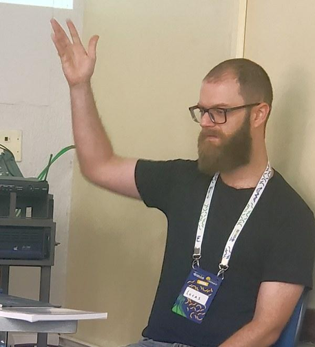 Foto de um homem branco de barba e óculos vestindo camiseta preta e crachá da Python Cerrado. Ele gesticula com a mão aberta erguida enquanto apresenta o seu tutorial.