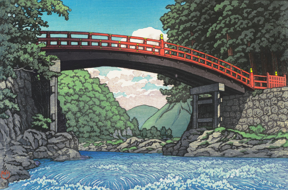 A view looking up at Nikko's Shinkyo Bridge on a bright summer's day. The red-lacquered bridge spans the Daiya River in a short, curving arc; tall trees crowd in close on either side. A view of the dramatic, mountainous terrain upstream is framed between the bridge and the river. Above, a few puffy white clouds populate an intense blue sky.