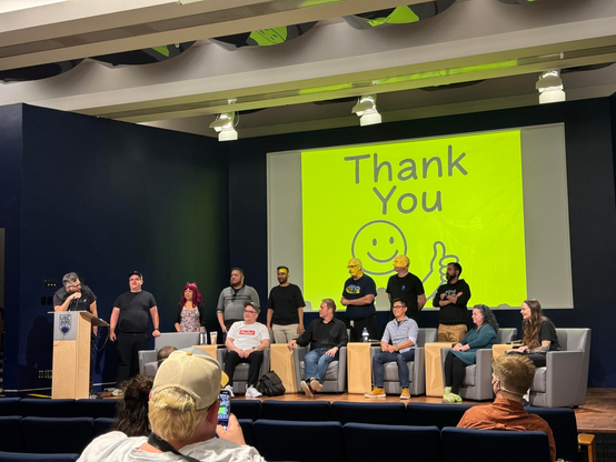 FediCon group photo, all speakers of the day standing in front of a yellow slide with a smiley face and a thumbs up that says "Thank You"