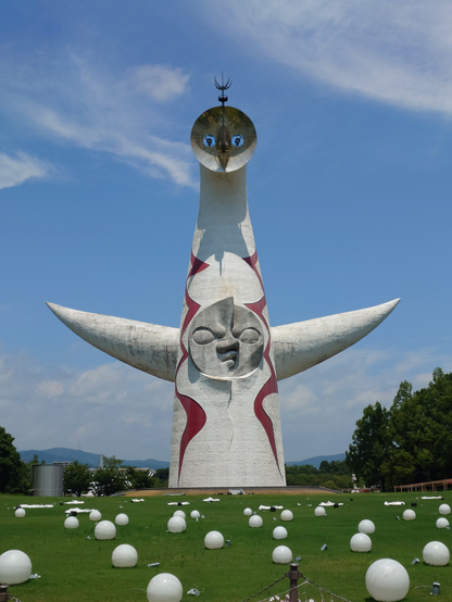 Taro Okamotos Sun Tower from afar standing in a park on a lawn. Basically a sculpture of a long white alien dude, with two pointy arms on each side, one angry looking face on its stomach and a golden round face on the top. 