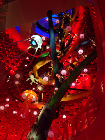 Inside the sun tower, looking at the tree of life from below. Very colorful lighting, red walls, a green tree, yellow and orange jellyfish and other simple lifeforms hanging from it. 