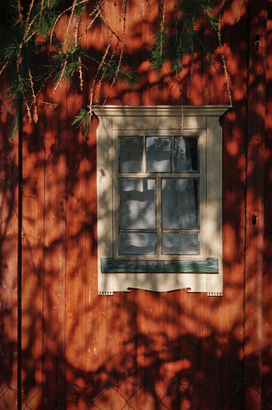 The photo shows a fragment of a wooden house with a window illuminated by the bright sun. A whimsical shadow falls on the wall through the branches of a tree, creating a play of light and shadow. The wooden wall of the house is painted in a rich terracotta color, while the window frame is a delicate cream shade.