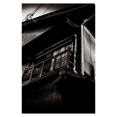 A monochrome photograph captures the upper section of an old wooden building in Nagoya, shot from a low angle that emphasizes its age and character. The balcony railings are worn and splintered, the sliding glass windows framed in dark wood. A metal drainpipe runs vertically along the side, patched in places, and electrical wires cut across the top corner of the image.