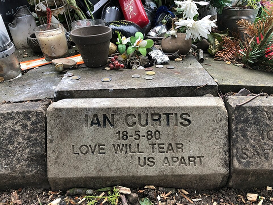 Small curb-like gravestone, engraved in a sans serif font with: 'Ian Curtis 18-5-80 Love Will Tear Us Apart'. Above it, on a flat surface, can be seen tributes, including flowers, candles, flowerpots, coins and a small metal skull. 