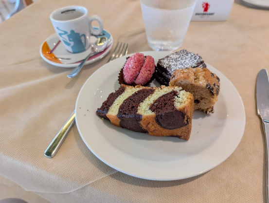 A high-angle close-up shot shows a white plate with four pastries on a beige tablecloth. In the foreground, a large slice of marble bundt cake with brown and yellow swirls fills the bottom half of the plate. To the left is a small pink cookie, and to the right is a small, light-brown cake square with visible chocolate chips. Behind that, a small, dark-brown brownie square is dusted with powdered sugar. A small white espresso cup with a light-blue design sits on a saucer to the left of the plate. A glass of water is in the top right, and a knife and fork are on either side of the plate.