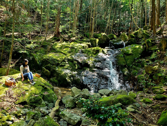 Nao-san relaxes beside Otowa-no-taki, the surrounding rocks carpeted in a lush green moss.