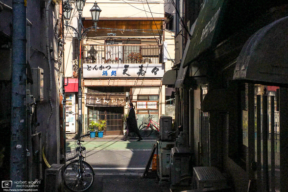 View of a Tonkatsu (fried pork cutlet) restaurant near JR Itabashi Station in Tokyo, Japan, seen through a narrow foreground alley of old buildings.
