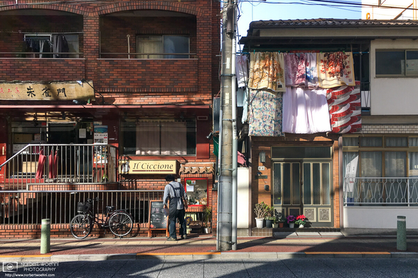 In the Oyama area of Itabashi Ward in Tokyo, Japan, a multitude of blankets and futons are hung out to air on the second floor of a buliding, atop its entrance. On the left side of the frame, a brick-style building is home to a few restaurants, outside one of which a man is checking the menu display boards.