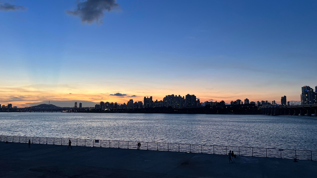 A sunset landscape focused on a city outline across a large river. The buildings are black against a sky turning from dark orange to dark blue. 