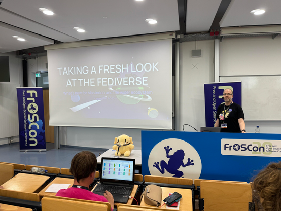 David Roetzel standing ready to present in front of a slide with the words “Taking a Fresh Look at The Fediverse”. A very cute, perfectly squidgy, beige, Mastodon mascot sits on a table, wearing a FrOSCon attendee badge.