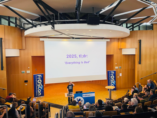 Andy Piper speaking at #FroOSCon in St. Augustin – and scratching his head. The slide above him says: "2025, tl;dr: Everything is bad."