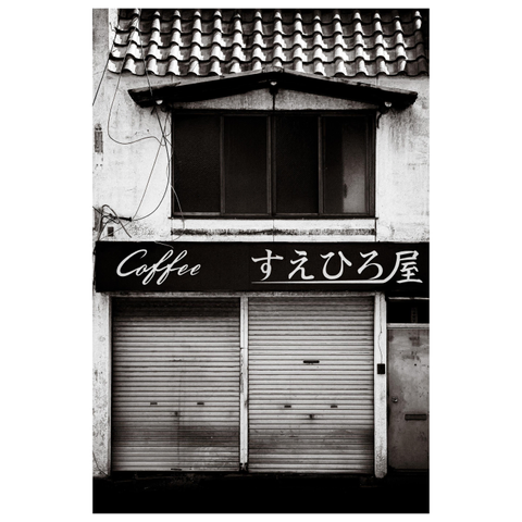 Description for the visually impaired: A black-and-white photo of an old Japanese coffee shop. The storefront has two closed metal shutters, above which is a sign reading “Coffee” in English and “すえひろ屋” in Japanese. The building shows signs of age with worn paint, tangled wires, and tiled roofing. The scene feels like a portrait of a café that once thrived but now sleeps behind its shutters.