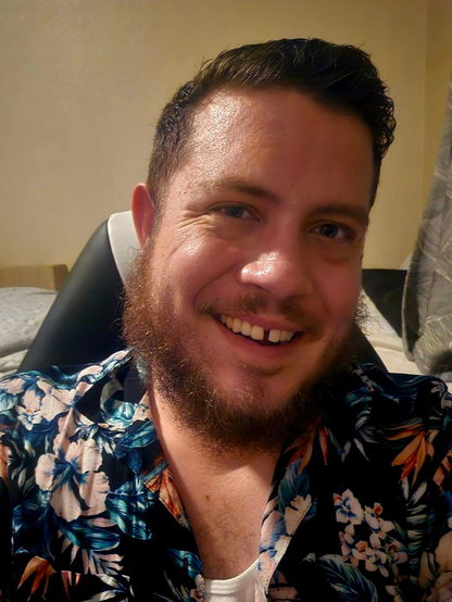 A picture of Sean, a 35-year-old white cisgender male with dark hair and a beard wearing a flowery shirt. His head is slightly tilted to the right and he's giving a toothy smile.