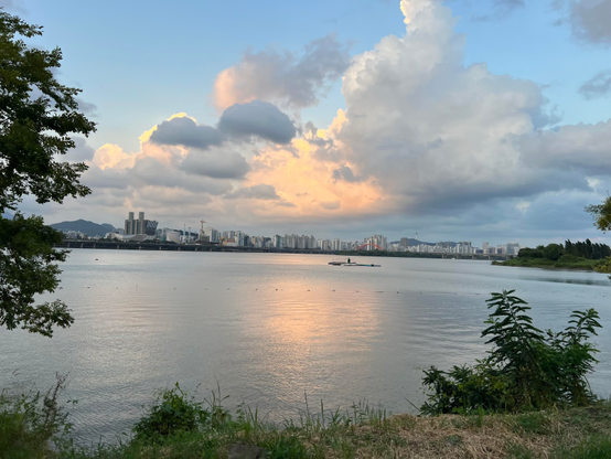A large river with a city skyline on the other bank and a small boat in the distance. The sky takes half of the picture and has big clouds with different shades of pink and grey. 