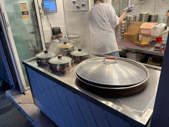 A steam-filled kitchen counter opened on the street with multiple steamed pots for dumplings. A person in a white shirt is working in the background, while kitchen utensils and containers are displayed around.