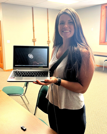 A smiling woman with long brown hair is holding a silver colored laptop on which the Linux Mint logo is visible.