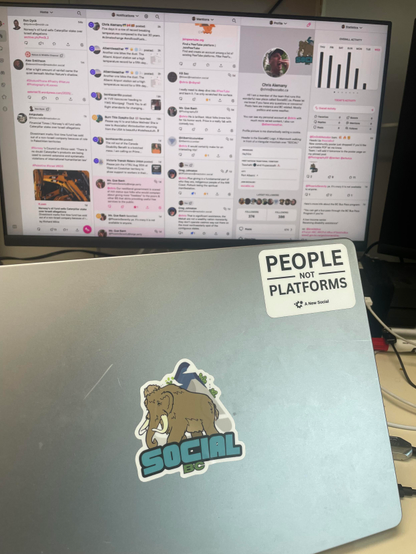 A picture of an aluminum laptop sitting on a desk with a flatscreen monitor in the background that is displaying my Ivory timelines. The laptop has a SocialBC logo sticker in the middle covering the Apple logo and there is a black-ink-on white sticker with the words “People not Platforms - A new Social”