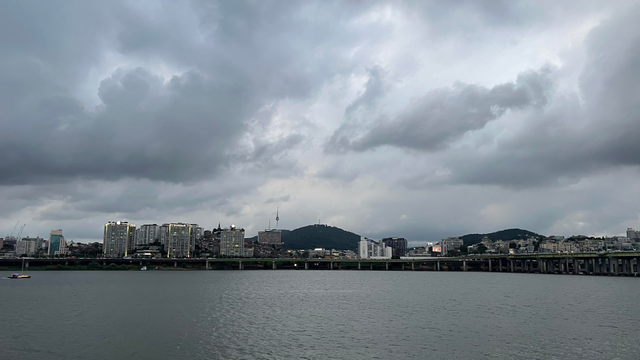 A view of a city skyline along a river, with tall buildings of different sizes and mountain ridge visible behind them and a thin tower standing on the mountain. The sky takes 2/3 of the picture and is filled with grey, menacing clouds. 