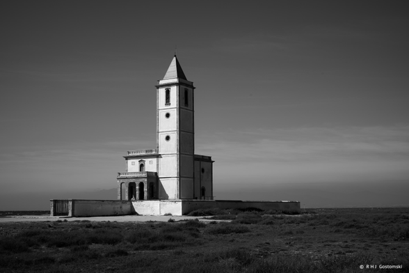 A black and white photo of a solitary church with a tall bell tower standing on a flat, grassy plain by the sea under a clear sky.
