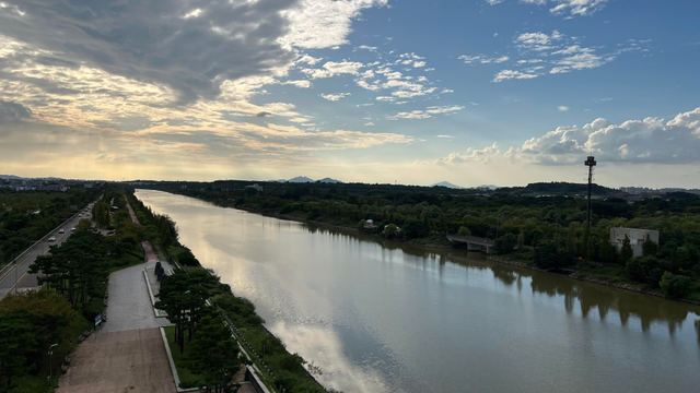 A scenic view of a river bordered by greenery seen from an elevated point. The sky is partly cloudy and it’s early evening. Sky and river are both light grey and blue which contrasts with the two banks being very dark. 