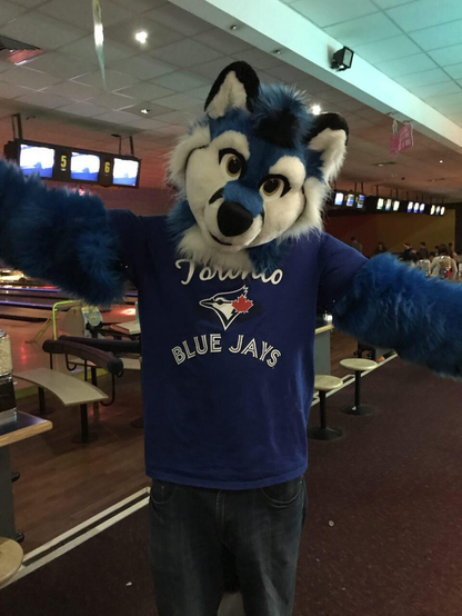 Blue fox partial fursuit, standing and with arms outstretched, at a bowling alley. He is wearing a Toronto Blue Jays t-shirt.