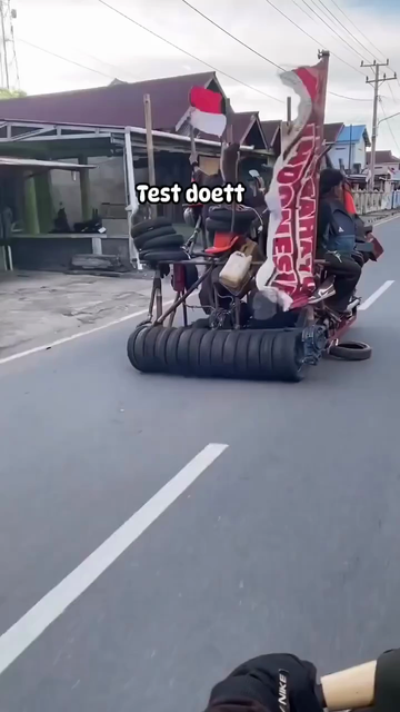 A bunch of people riding a makeshift platform made out of bamboo sticks on a road, powered by a couple of motors, steered with a welded tiller. A bunch of small, thin spare tires are installed all over the contraption, and two monkeys are clambering all over. The vehicle displays the Indonesian flag and a banner celebrating their independence day.