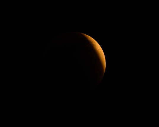 A partially illuminated view of the moon against a black background, featuring a reddish-orange hue during the lunar eclipse.