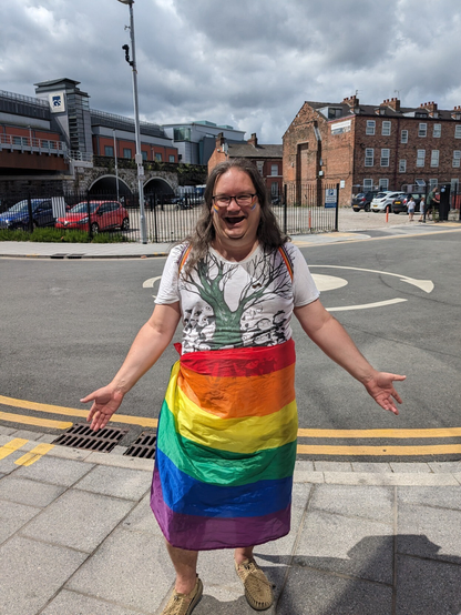 Me. At Warrington Pride at the end of June 2023. Sunny, outdoors with a mini roundabout behind. I am wearing a rainbow flag (not the progress pride flag, sadly) round my waist and a white t-shirt with a tree and Moomin-like creatures on. I have long, un-styled hair and glasses. I have a rather over the top goofy smile.