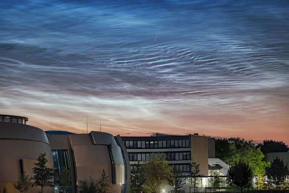 Blue wavy clouds – almost like the surface of a pond – above a group of buildings. At the top-centre of the image there's a small white dot with an short tail: a comet.