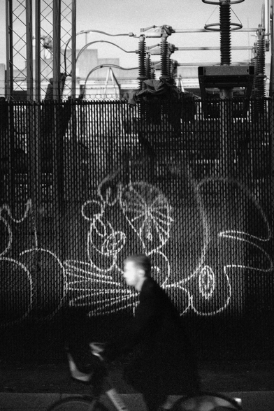 A man rides a bicycle into a spot of light past an electrical substation.