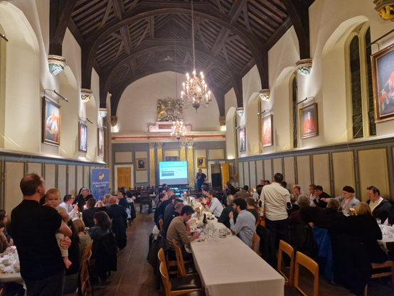 Thomas presenting his Feuding Pythonistas game in a grand hall to three long tables of the core team and friends.
