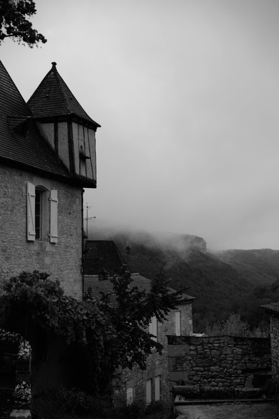 Black and white photo of a stone building with a pointed turret and shuttered window in a foggy, mountainous village