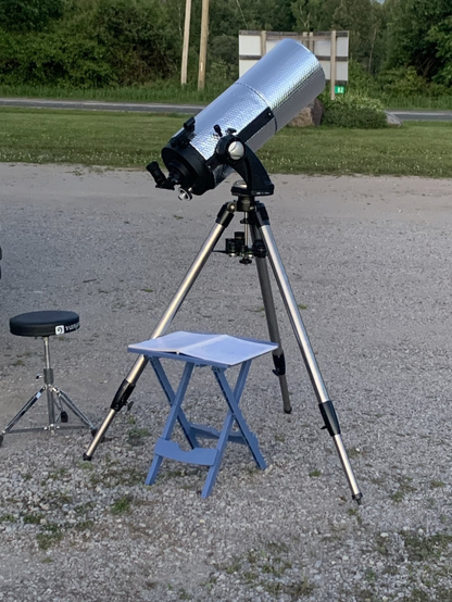 A picture of my telescope (a 1985 Meade 2080 LX3) sitting in a gravel parking area with my drum throne and a small table beside it. On the table are some paper star charts (paper like a pirate! Arrrr!).