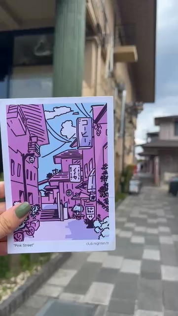 Video: A hand holding a print of an illustration showing a Japanese street in pink shades. The hand then move over, revealing the exact street that was used as a model