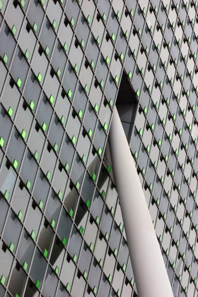 The facade of a tall office building, photographed diagonally from below. A play of lines emerges from wide rows of square glass windows, alternating dark and light gray, from the upper left to the right, while diagonal, narrow lines also run from the lower left to the upper right (of the window jambs). On each window is a small, square lamp, projecting, creating a shadow in the window. The lamps emit green light. From the lower right, a large white column disappears into a hole in the middle of the building. The column is reflected in the glass wall.