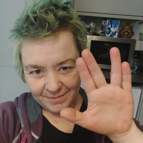 Me, a white woman with very short, faded dyed green hair giving a Vulcan salute to camera. I have a purple and grey striped hoody, a black t shirt and am sitting in front of some grey cupboards and a microwave.