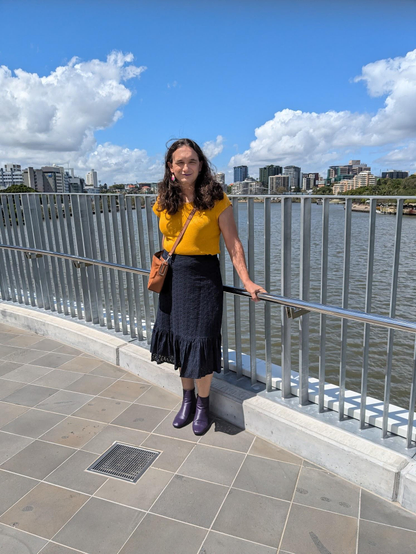 Davie, wearing a yellow short sleeved top, a navy broderie skirt, purple boots and with a brown cross body bag. Standing in front of a hand rail on the Neville Bonner Bridge with Meanjin/Brisbane and the river in the background.