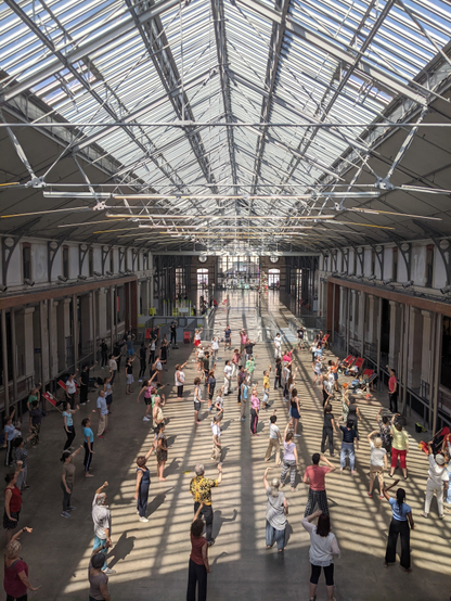 Grande Nef ensoleillé du CENTQUATRE-PARIS avec des personnes pratiquant du qi-gong et levant toutes et tous la main sous la verrière