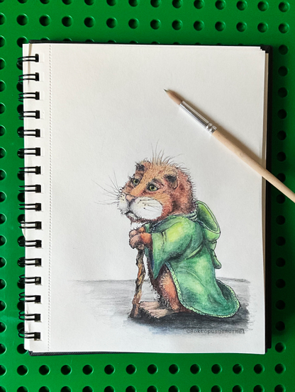 Blick auf ein Skizzenbuch, in dem ein aufrecht stehender Hamster in grünem Kapuzenmantel zu sehen ist. Er stützt sich mit beiden Vorderpfoten auf einen knorrigen Gehstock.