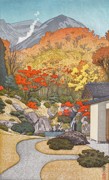 A bright autumn day in the gardens at Hakone Museum. In the foreground, a narrow path curves and divides: one branch leads toward an archway on the right, while the other leads toward a small pond enclosed by a steep banks up ahead. Trees and shrubs of various kinds populate the landscape, all turning yellow and orange and red with the season. Beyond, dramatic mountains dominate the view; a trail of white smoke rises from a valley somewhere among them, drifting off into the clear blue sky.
