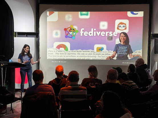 A woman is speaking at a presentation about the "fediverse," with a colorful slide behind her featuring various social media icons and text. The audience, sitting in rows, is focused on her as she discusses decentralized social networks.