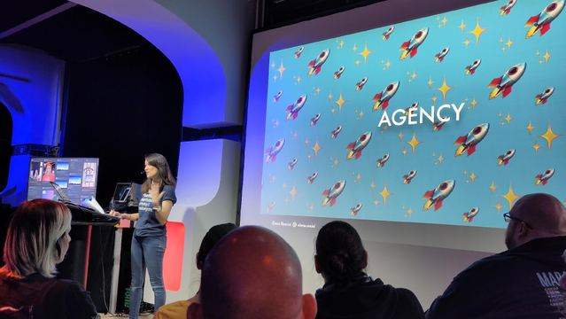 Woman in vor T-Shirt in a hall in front of people, background presentation saying "Agency"