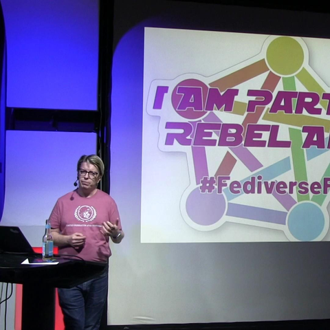 Andy wearing a pinkish Fediverse T-Shirt at Fediday. in the background there is a slide showing his Fediverse Sticker.