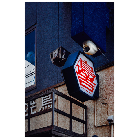 A color photo showing a glowing hexagonal shop sign with a red kanji character at its center. The sign juts out from a textured wall painted dark blue and beige. Above and beside it, a security camera and light fixtures echo the angular shapes.
