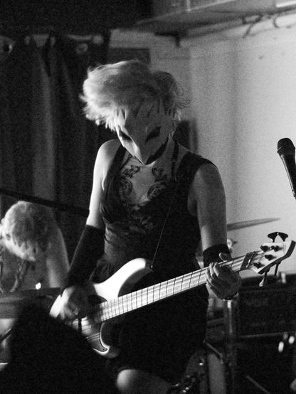 A black-and-white photo of a girl with short hair on a stage playing bass guitar. She is wearing a black dress and a white birdlike mask. Photo by Orlando daSilva