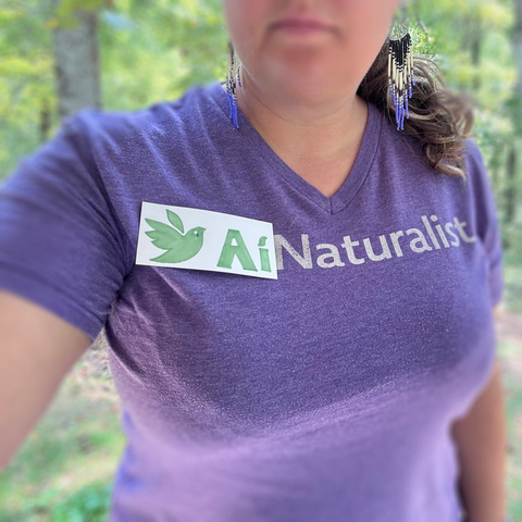 A person takes a selfie with the background blurred forest. The shirt was a purple shirt with the iNaturalist logo and text, and has been altered with a hand drawn logo to add an "A" before the the "i" in iNaturalist, so it now reads "AiNaturalist" in protest. The iNaturalist bird has a tear under the eye as we are saddened by the looming death of integrity.